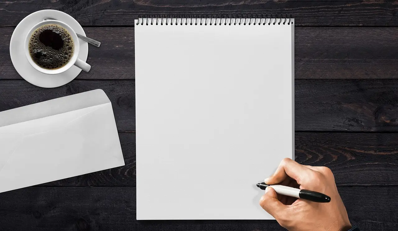 A blank spiral notepad, a coffee cup, one envelope and a hand holding a marker